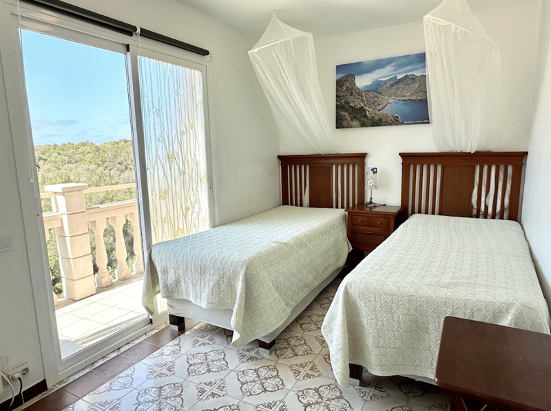 Two twin beds, wooden headboards, nightstands, patterned tile floor, large window with balcony, artwork on the wall