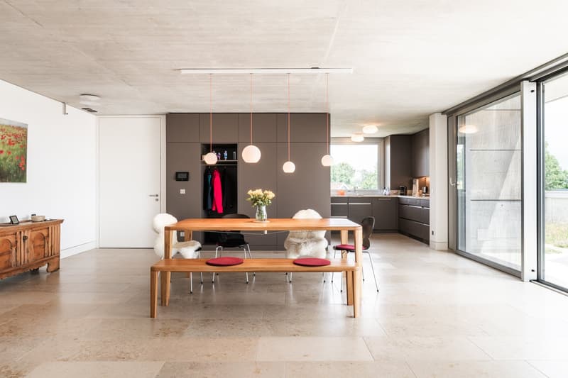 Modern kitchen with large dining table and bench, glass doors, wooden cabinet, flowers, white walls, tiled floor.