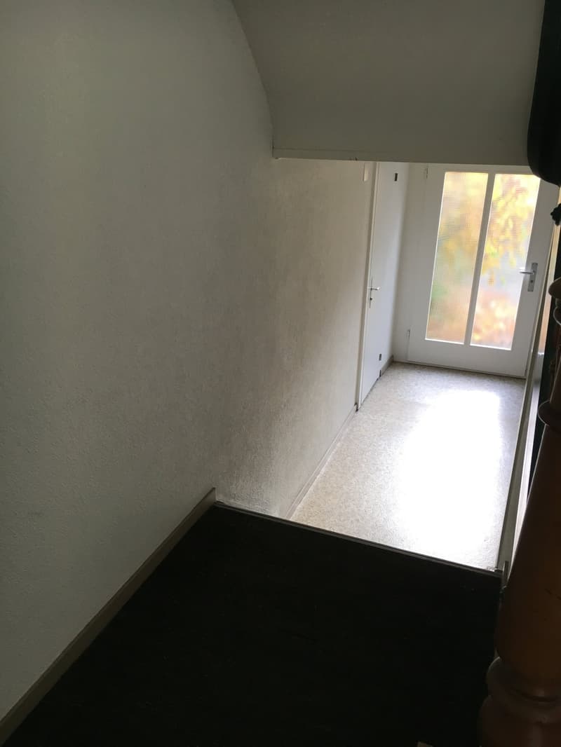 The image shows a small, empty room with white walls and a window. The floor appears to be covered with a dark-colored material, possibly a carpet or linoleum. The room has a simple, minimalist design with no visible furniture or decor.