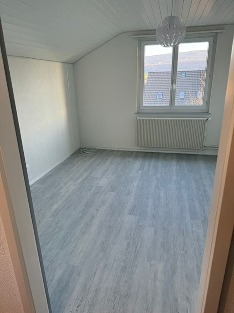 Empty room, white walls, wooden-look flooring, central heating radiator, window overlooking buildings.