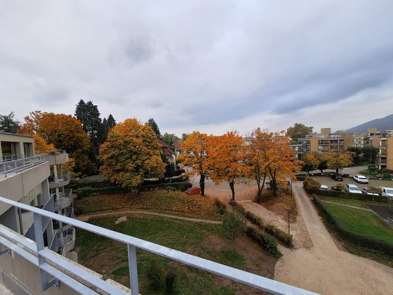 balcony with railings, multiple trees, green grass, concrete pathway, parking area with parked cars, residential buildings, cloudy sky