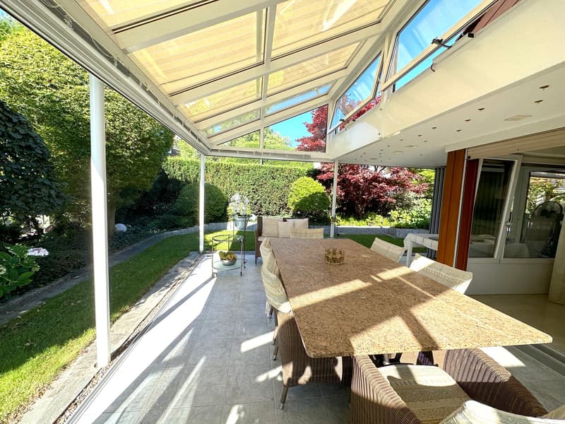Outdoor dining area with a large table and chairs, glass walls, and a metal frame. The area is covered with a white awning and has a tiled floor.