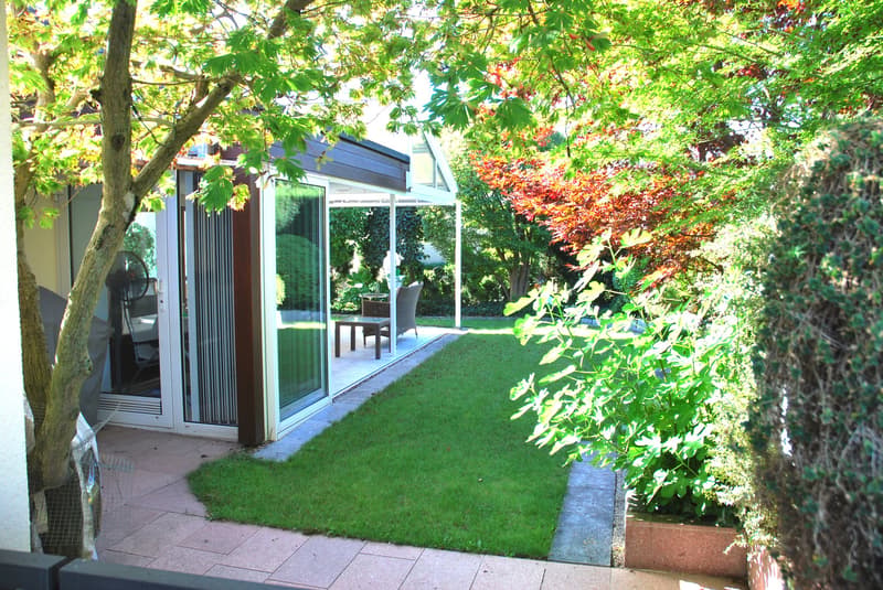 A garden with a patio featuring a glass door leading to the interior, a grassy area, and a small table with chairs under a canopy. There are trees and bushes surrounding the garden, and a pathway leading to the patio.