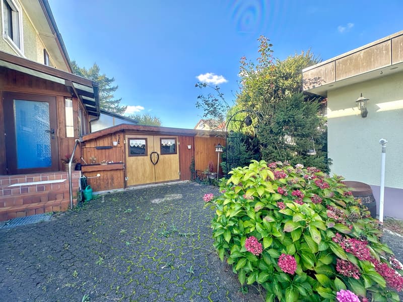 Backyard with brick paving, wooden shed, flowers, wooden fence, another house at the end of yard.
