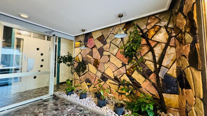 Tiled floor, mosaic wall with plants, glass doors, ceiling with hanging lamps, windows with grills.