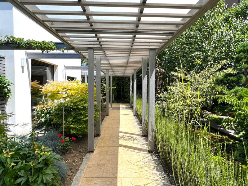 A pergola walkway with tiled flooring in a garden, surrounded by various plants and trees, leading towards a house.
