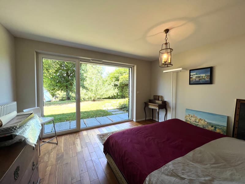 Sliding glass doors leading to a garden, wooden floors, large windows, white walls, two paintings on the wall, bed, chair, dresser, table, chandelier.