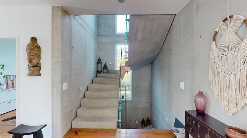 A modern house with a raised ground floor, stone stairs, glass railings, concrete walls, and a wooden floor. The living area has a glass window, a Buddha statue, and a white wall.