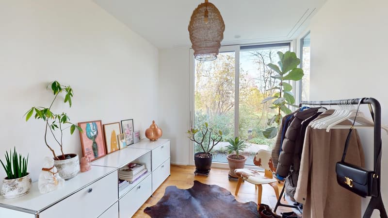 White walls, white cabinet with figurines, plants, modern chair, rug, hanging clothes, picture frames