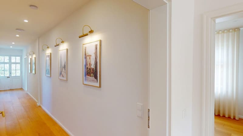Long hallway with wooden floors, white walls, framed art pieces, gold light fixtures, and a white door.