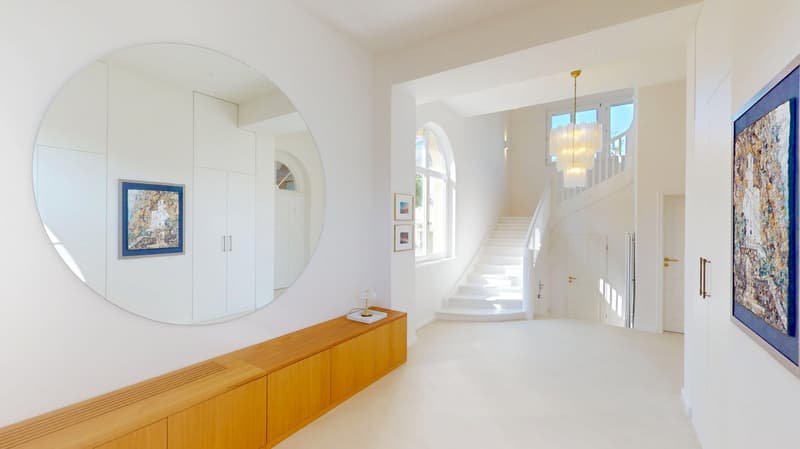 A hallway with a large round mirror on the left side and a wooden bench. A chandelier is lit above the staircase on the right. White walls and flooring with two pictures and an arched window on the right.