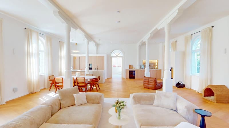 Modern open plan living space with wooden floor, white walls, dining table and chairs, beige couches, kitchen with bar counter and windows.