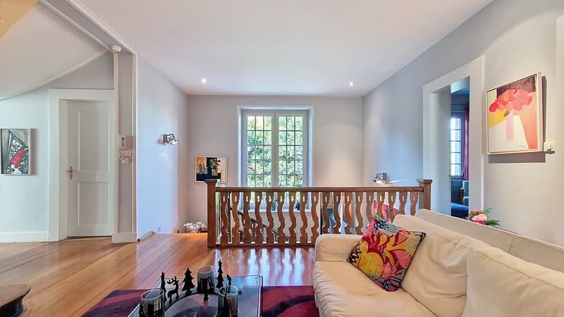 Modern interior with wooden floor, white walls, stairs, and decorative elements. Features include sofa, table with candles, paintings, and decorative items.