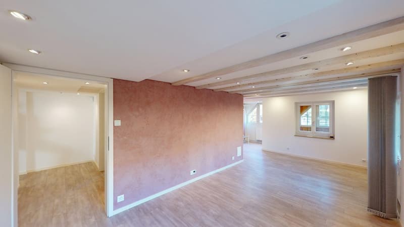 Empty room with wooden flooring, white walls, one pink accent wall with two power outlets, ceiling with wooden beams and recessed lighting, open door to another room.