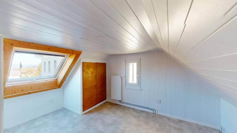 A corner room with wooden windows, wooden ceiling beams, a door, radiator, wooden flooring, and white walls.