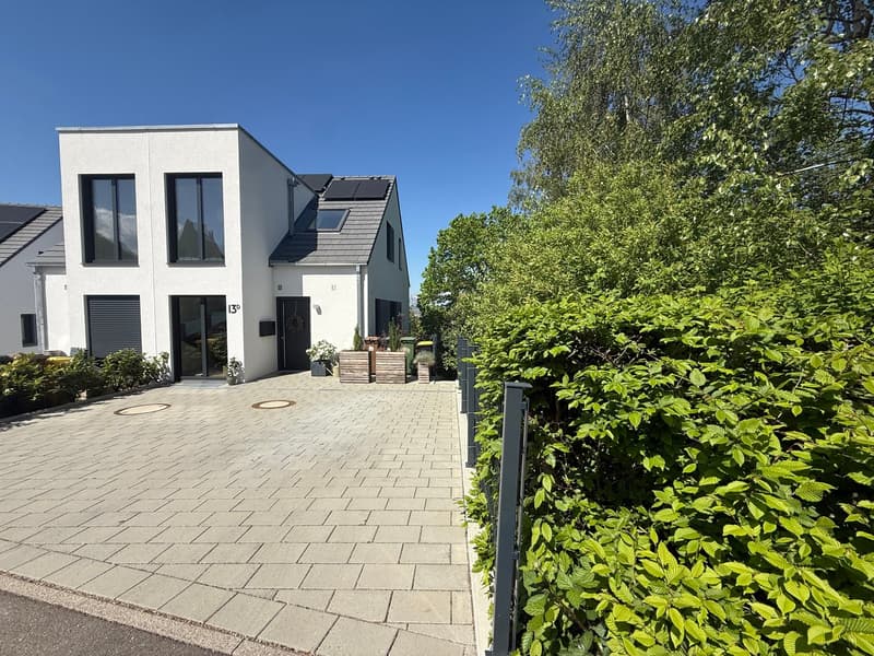 Modern white house, solar panels on the roof, brick driveway, shrubs and trees, trash bins