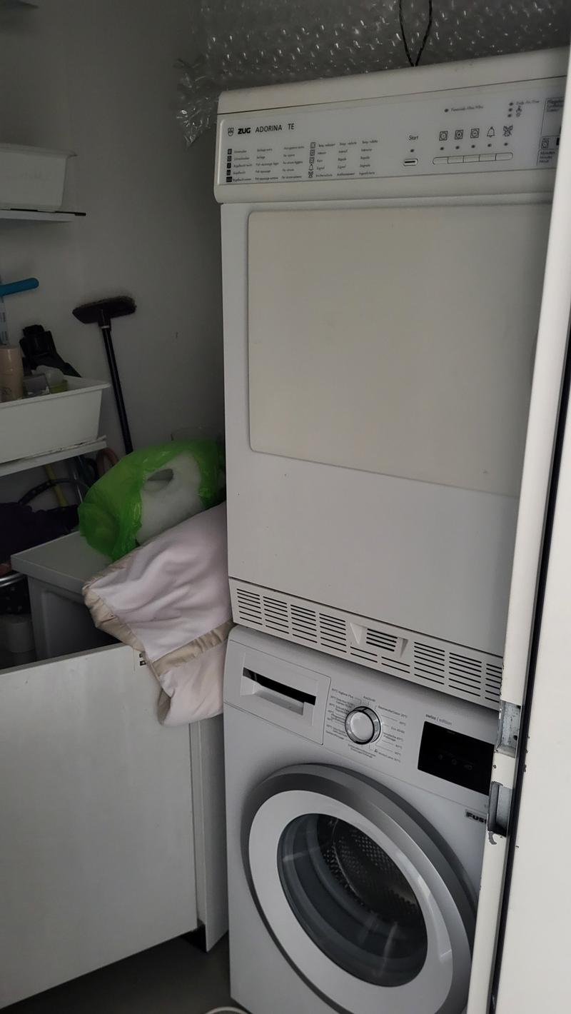 A room with a washing machine and a dishwasher stacked on top of each other, a white cabinet, and a white shelf with various items on it.