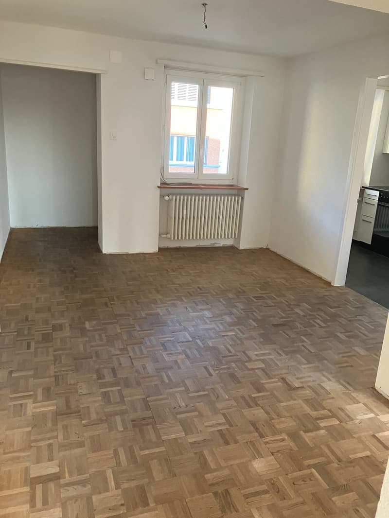 Empty room with wooden parquet flooring, white walls, a window, and a radiator below it. A door leading to another room is on the left side, while an open door with a kitchen glimpse is on the right side.