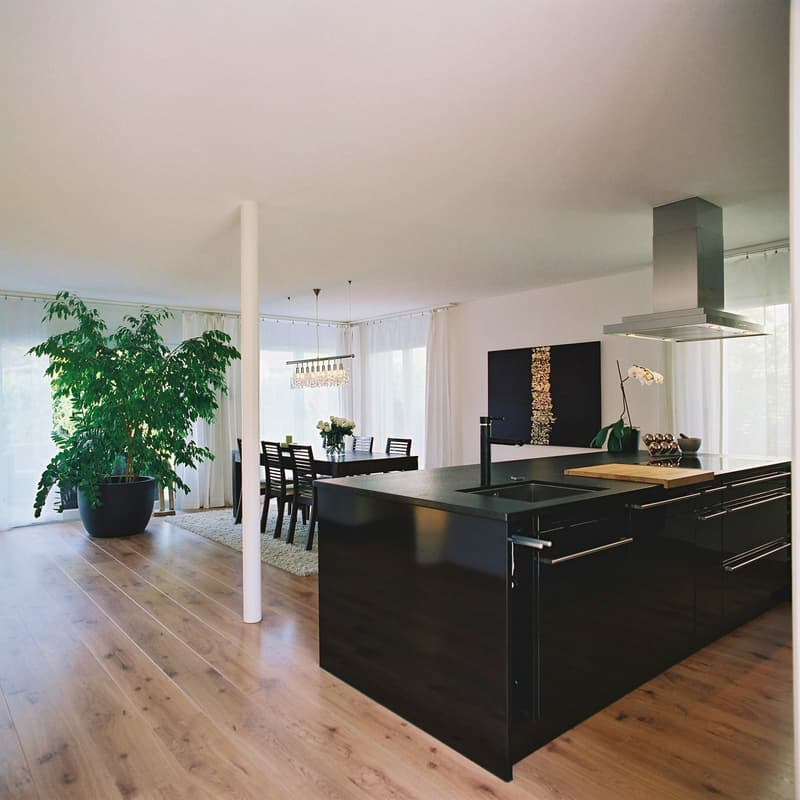 Open plan kitchen and dining room with wooden floor, chandelier lighting, modern black island with marble countertop, kitchen sink, gas range, and dining table with chairs