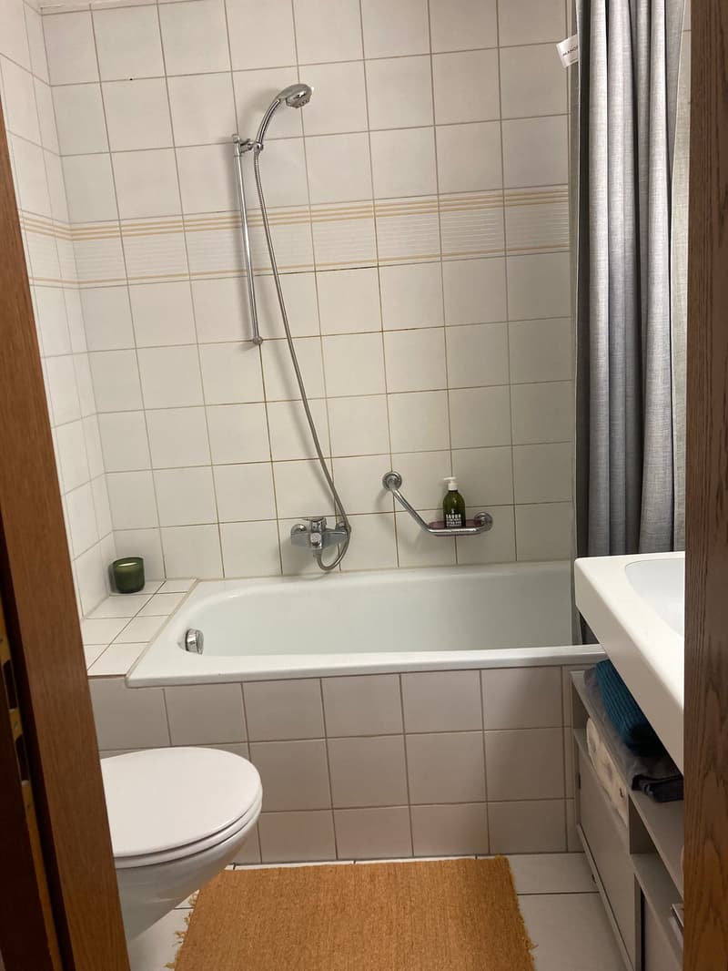 White tiled walls, white bathtub, white toilet, wooden rug, silver shower head