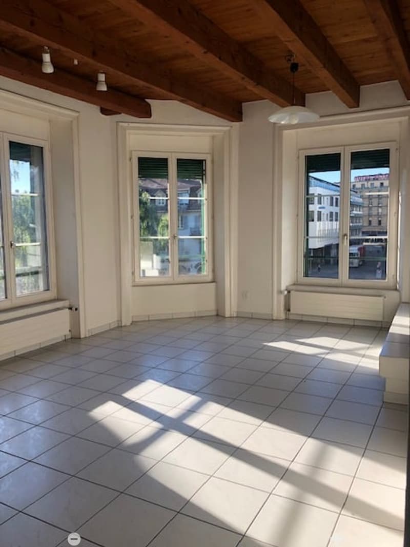 Empty room with white tiles, wooden beams, windows, and a ceiling lamp