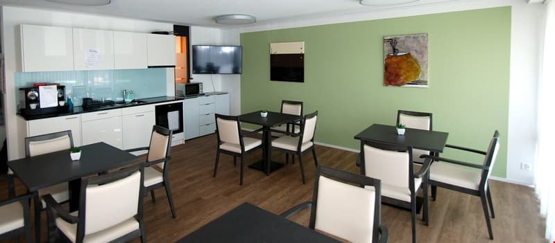 Dining area with tables and chairs, green wall, white cabinets, kitchen appliances, paintings