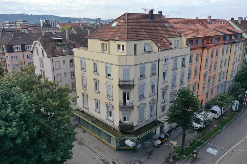 Aerial view of a multi-story building with brown roofs and multiple windows. The building is surrounded by other buildings and trees. There are vehicles parked on the side of the road.