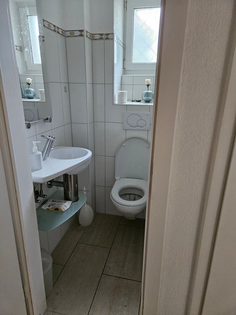 A small bathroom with a sink, toilet, and window. It has white tiles, a mirror, and a small flower vase. The sink has a faucet and a soap dispenser. The toilet is white and has a toilet paper holder. There is a toilet paper roll on the wall. The bathroom