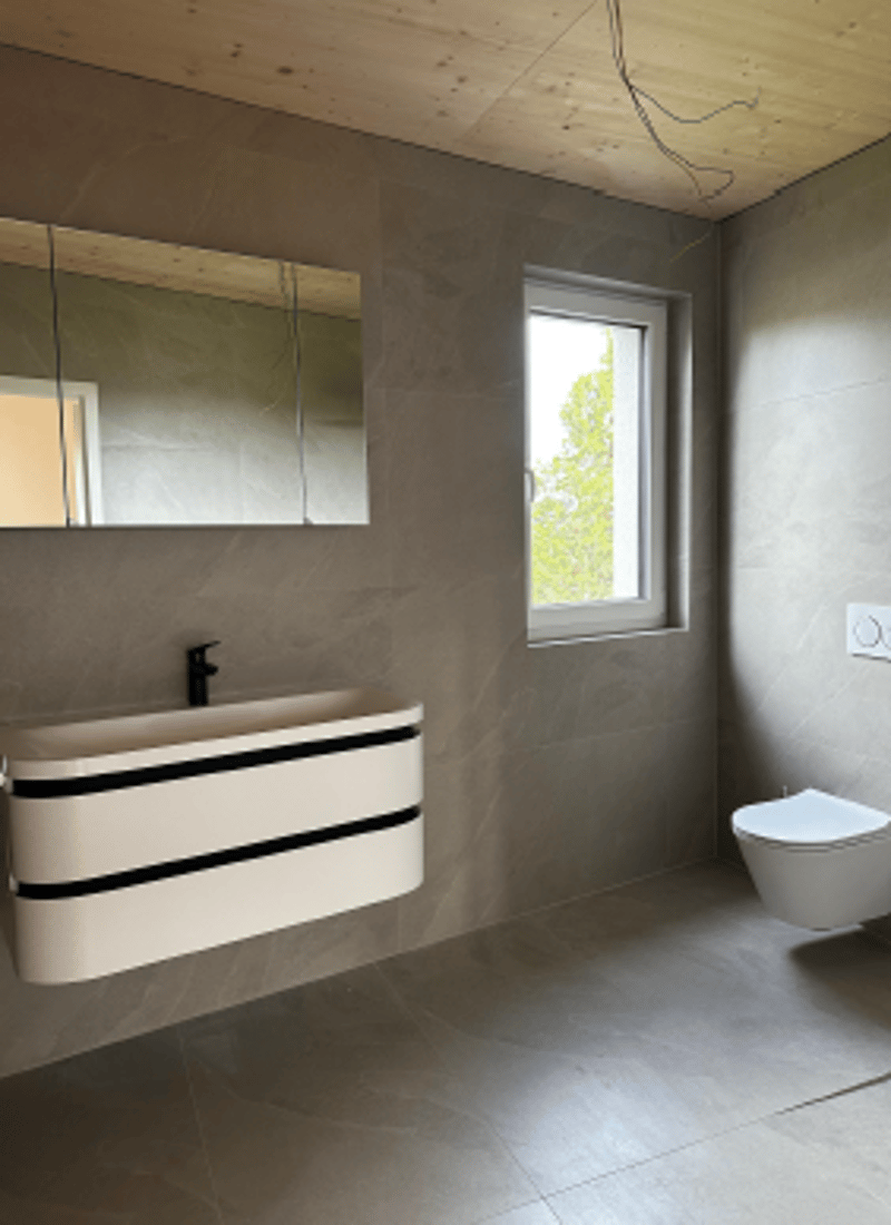 Modern bathroom with a white vanity, mirror, and toilet. Gray tiled walls and floor.
