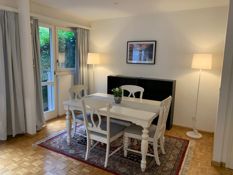 Bright and airy dining room with large windows, white wooden dining table and chairs, black sideboard, framed artwork on the wall, and a decorative rug on the hardwood floor.