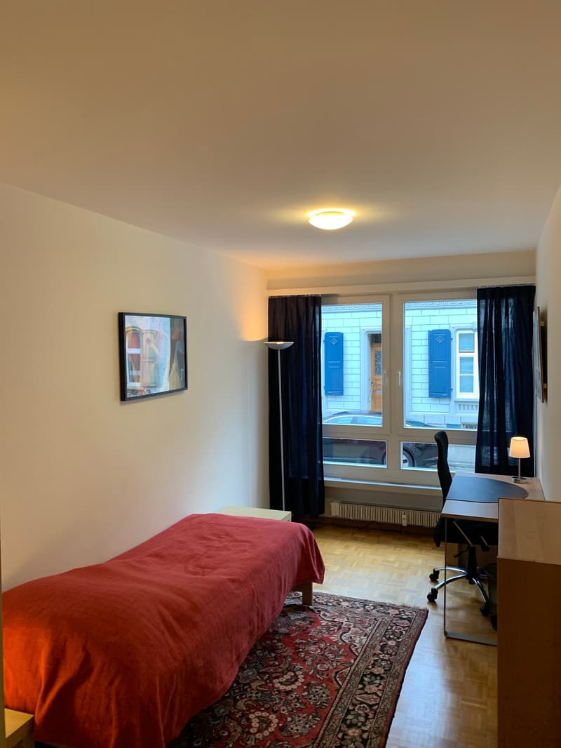 Bedroom with a red bedspread, a desk with a chair, and a framed artwork on the wall. Large windows with blue shutters provide natural light and a view of the exterior.