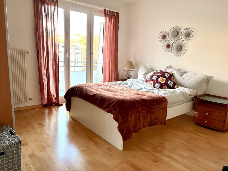 bedroom with white bed, red bedspread, white pillows, lamp, side table, bedside drawers, hardwood floors, balcony