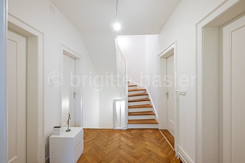 Bright and spacious hallway with hardwood herringbone flooring, white walls, and a staircase leading upstairs. The hallway has multiple doorways leading to other rooms.