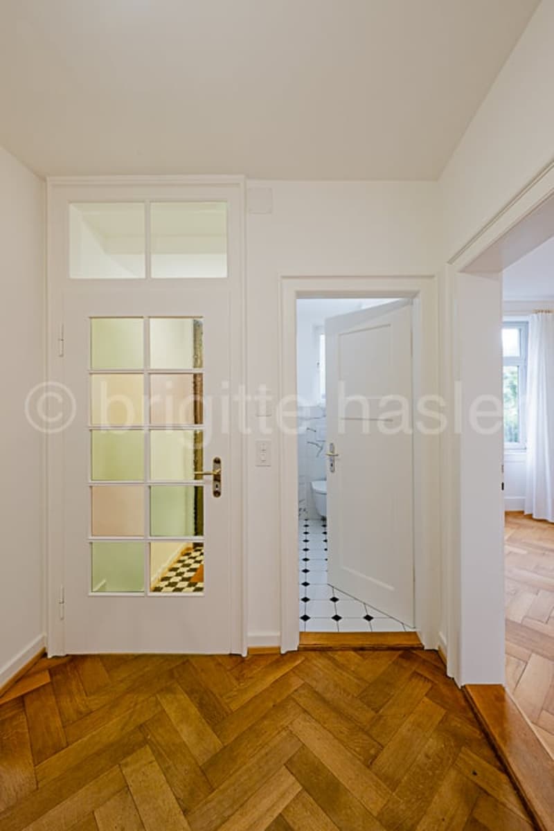 Bright and spacious hallway with white walls, wooden herringbone flooring, and a glass-paneled door leading to what appears to be a balcony or terrace.