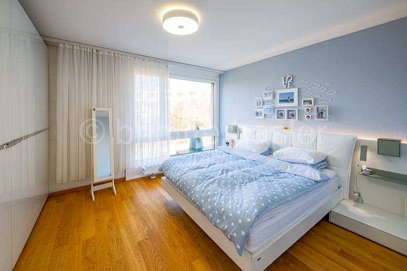 White bed, blue polka dot bedspread, multiple pillows, bedside tables, mirror, white curtains, wood floor, light fixtures