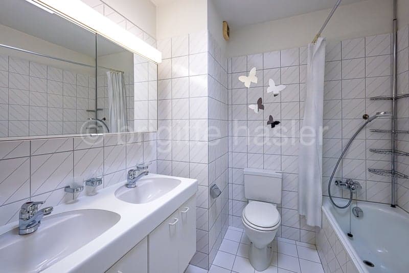 Bathroom with white tiles, double sink, mirror, toilet, bathtub, shower curtain, and wall-mounted shelves