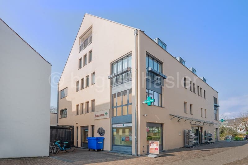 A beige building with glass windows and a green sign on the wall. There is a bicycle parked in front of the building and a blue trash bin on the left side. There is a signboard on the right side of the building.