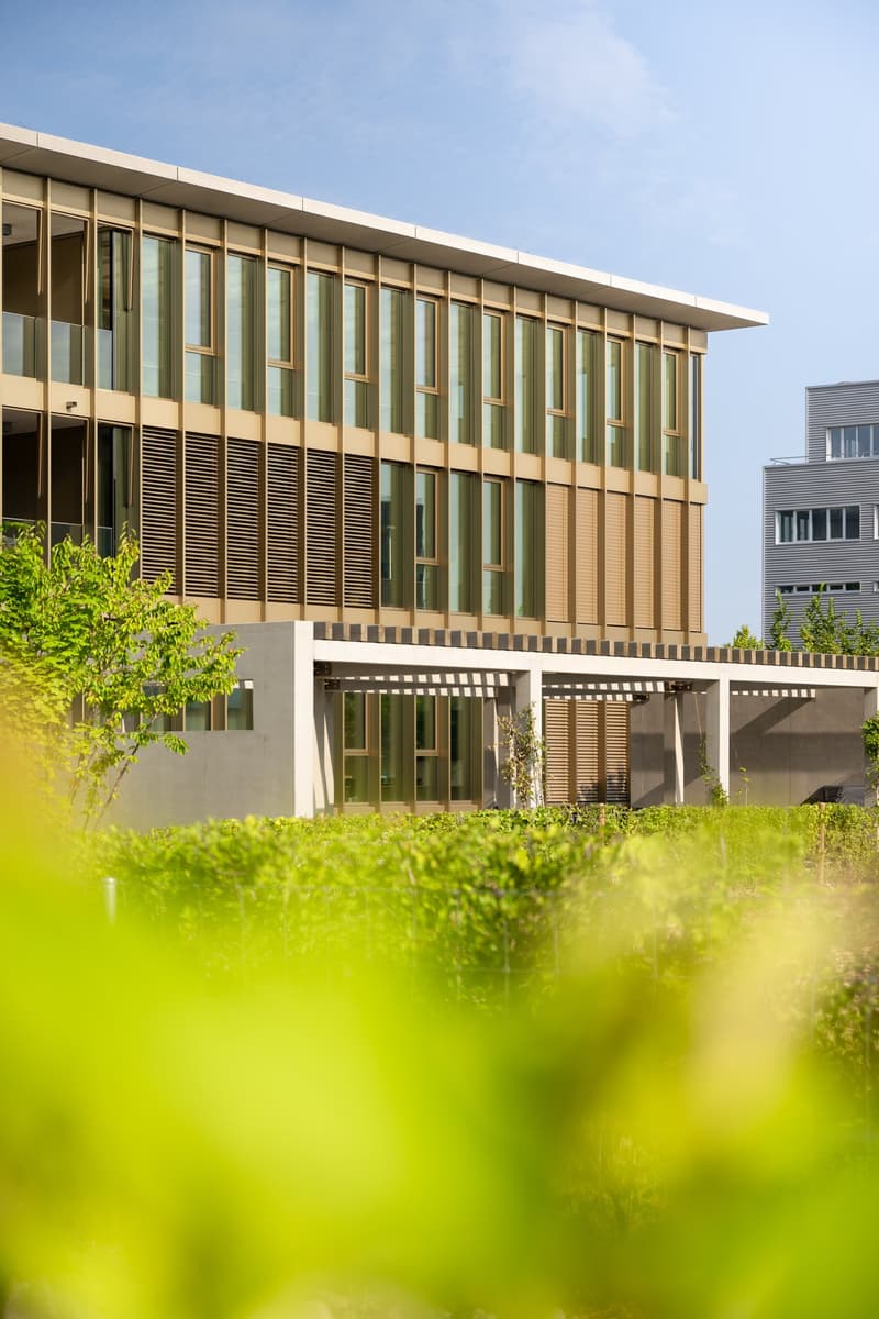 Multi-story office building with a modern, glass-and-concrete facade. The building has rows of large windows and a covered entryway. The surrounding area is landscaped with greenery.