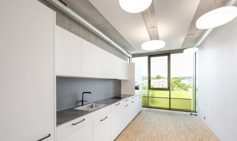 Modern, open-concept kitchen with white cabinets, stainless steel sink, and large windows overlooking a balcony or terrace with a view of the surrounding greenery.