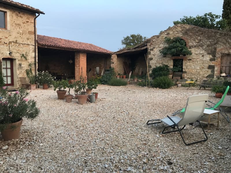 Two-story stone house with a covered patio, gravel courtyard, potted plants, and a reclining chair