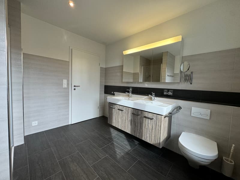 modern bathroom with a dual sink, mirror, toilet, and power outlet. Tiled floor and wall with black border.