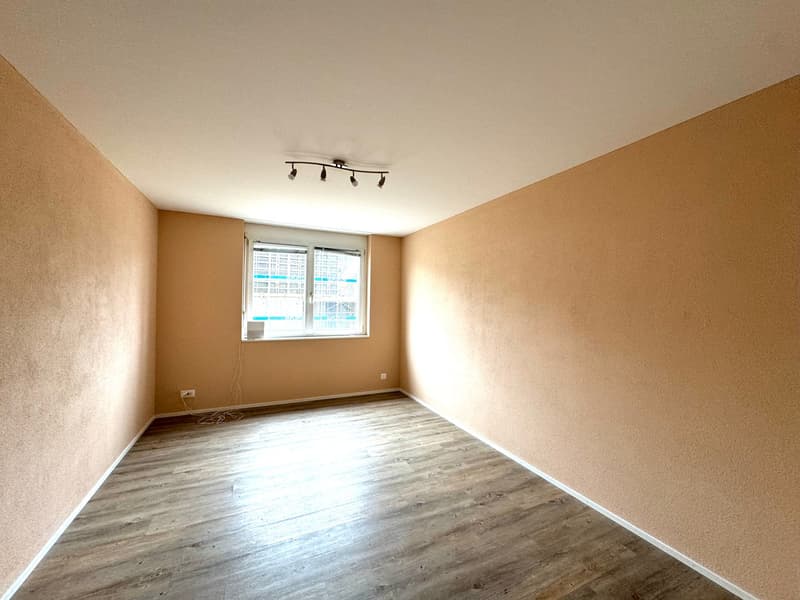 Empty room with wooden floor, beige walls, ceiling lights, window, power outlets.