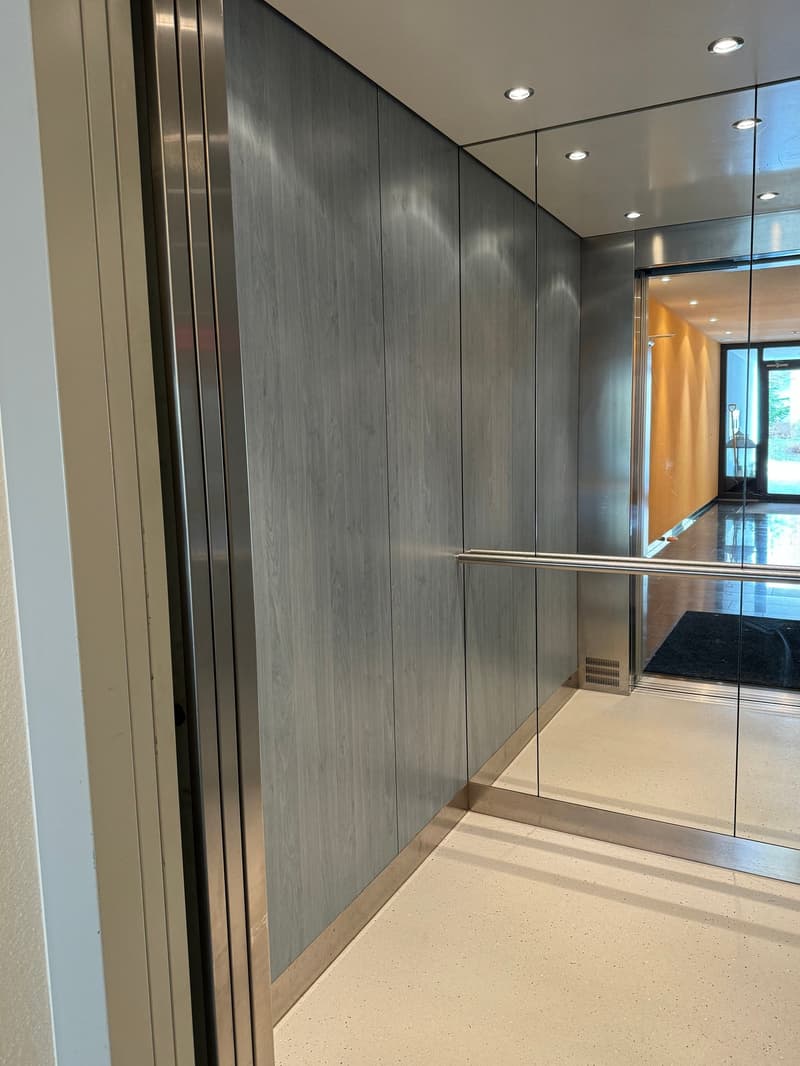 Modern elevator, wood-like panels, metallic details, mirror-finish surfaces, high ceilings with lighting, sleek flooring, interior doors leading to rooms