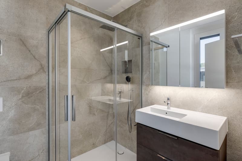 Large walk-in shower with glass enclosure, white vanity with sink, and large mirror above the vanity. The bathroom has a modern, minimalist design with gray marble-like tiles covering the walls.