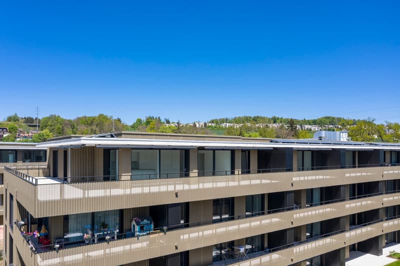 Multi-story apartment building with balconies, surrounded by trees and greenery, under a clear blue sky