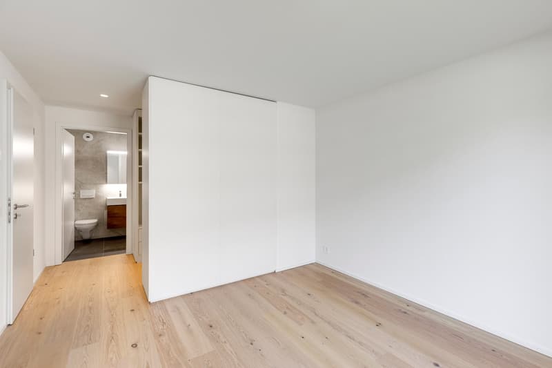 Spacious bedroom with white walls, hardwood flooring, and a large closet. The bathroom is visible through the open doorway.