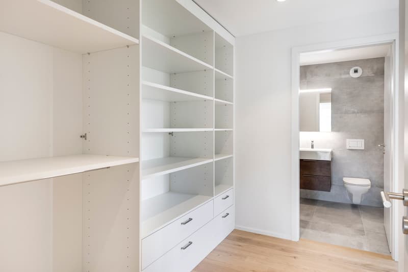 Spacious bedroom with built-in white shelving and drawers, hardwood flooring, and an adjoining bathroom with a modern vanity and tile flooring.