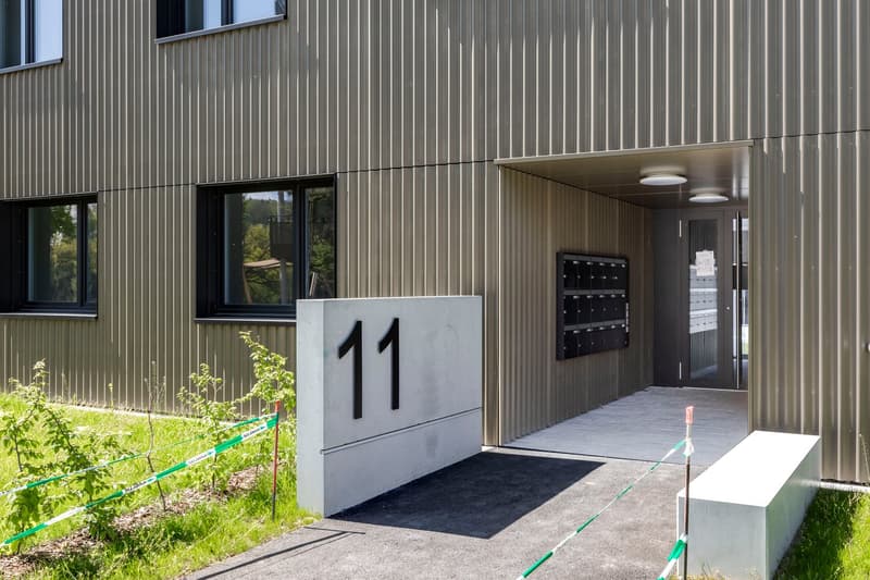 The image shows the exterior of a modern, industrial-style building with a gray metal facade. The building has large windows and a numbered entrance, indicating it is likely an apartment or office building. There is a small garden area with plants in the
