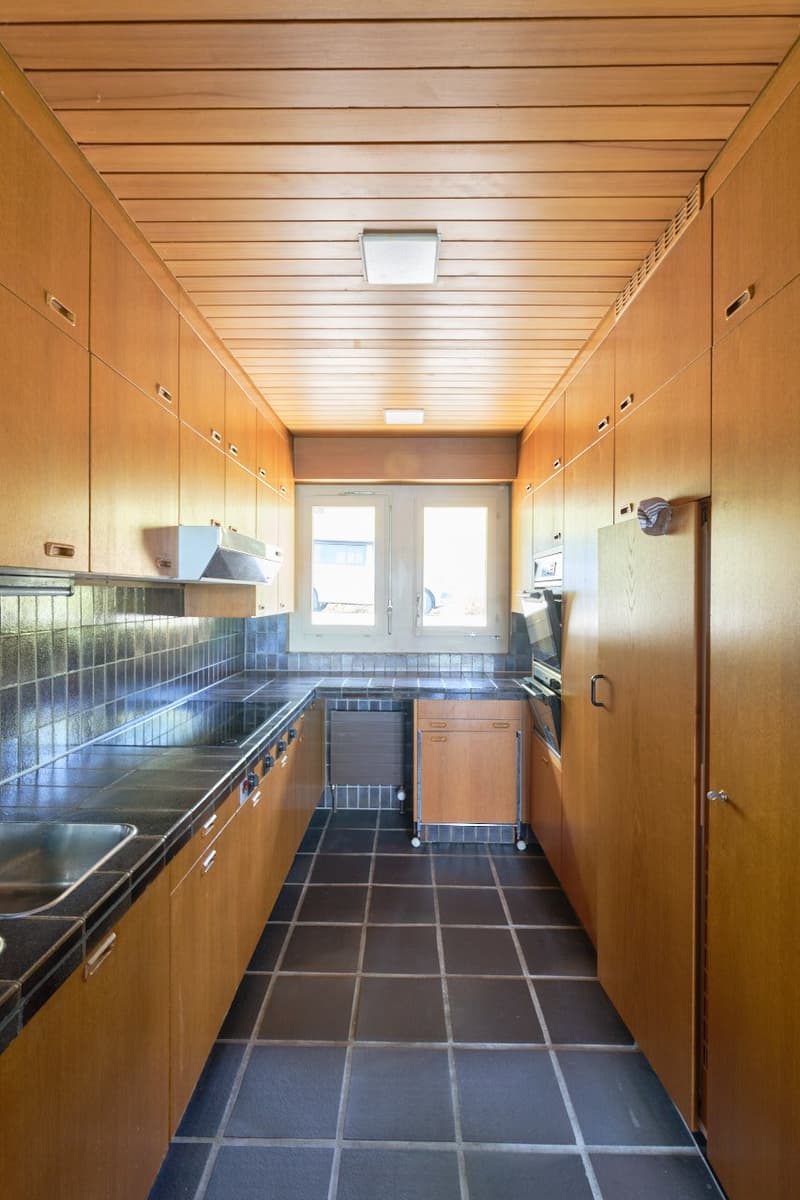 Galley-style kitchen with wooden cabinets and shelves, tiled floor, stainless steel appliances including a dishwasher and washing machine, and a window providing natural light.