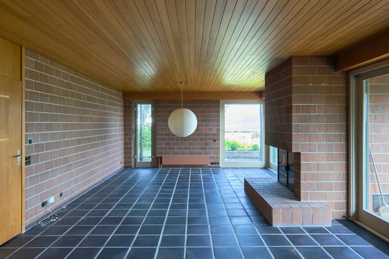 Large open-plan living space with brick walls, wooden ceiling, tiled flooring, and a fireplace feature. The room has large windows providing natural light and views of the outdoor area.